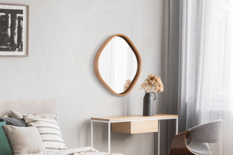 Elegant round mirror in wooden frame above fancy console table with flowers in vase in trendy bedroom interior with beige vase